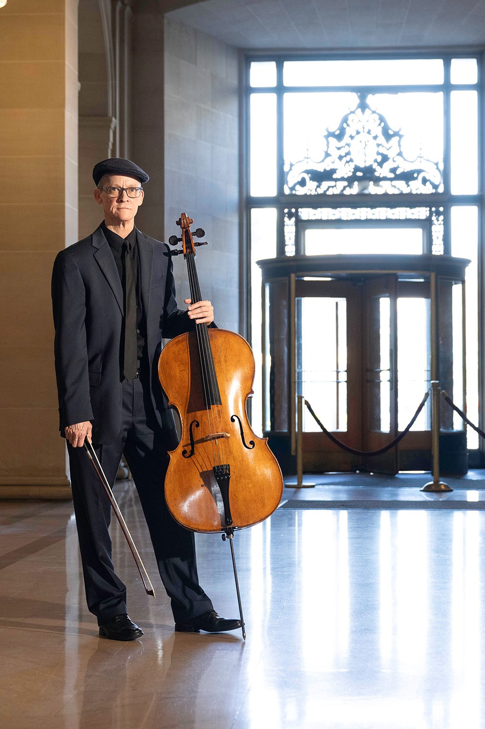 Ben Welch Snellings in formal attire stands with a cello and bow in an elegant lobby, showcasing a blend of artistry and architectural beauty.