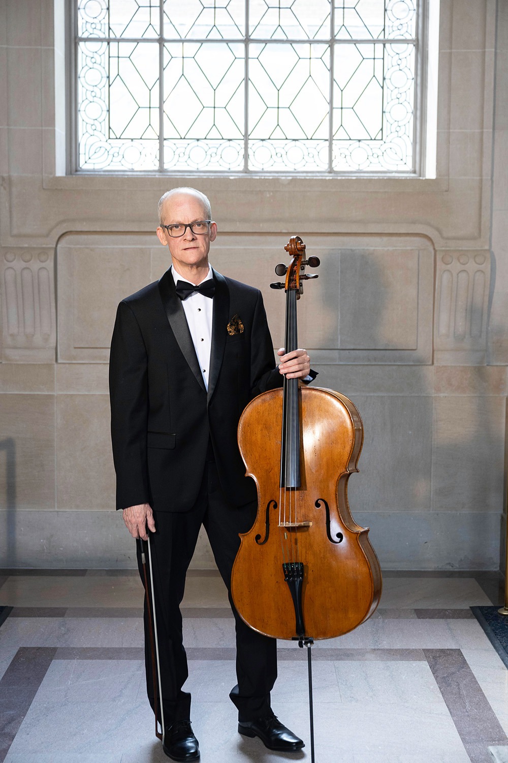 Ben Welch Snellings in a tuxedo stands with a vintage cello, posing in front of an elegant stained glass window, showcasing a blend of music and art.