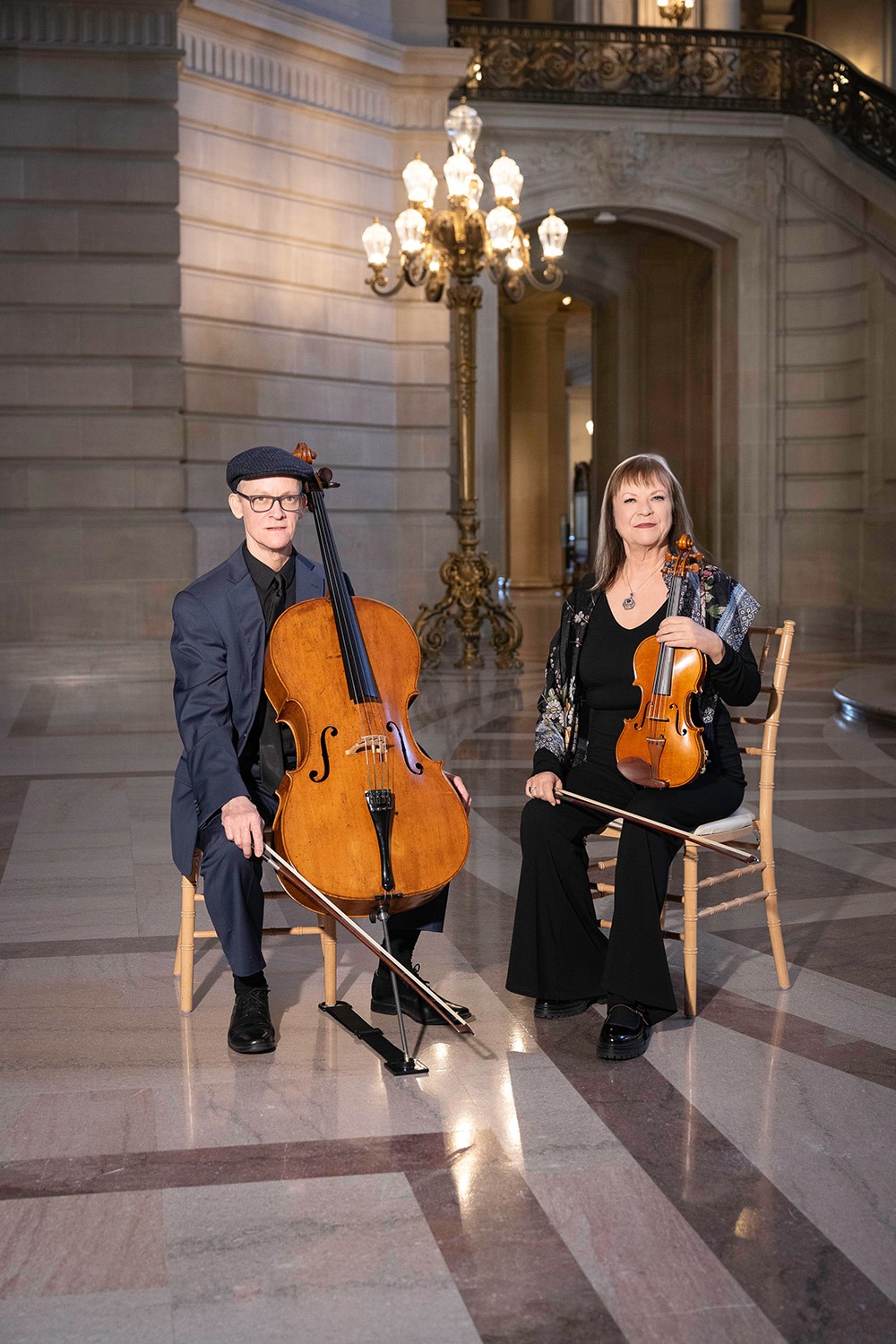 A cellist in a suit and a violinist in black attire pose in an elegant hall, showcasing their instruments. The setting emphasizes a musical performance.