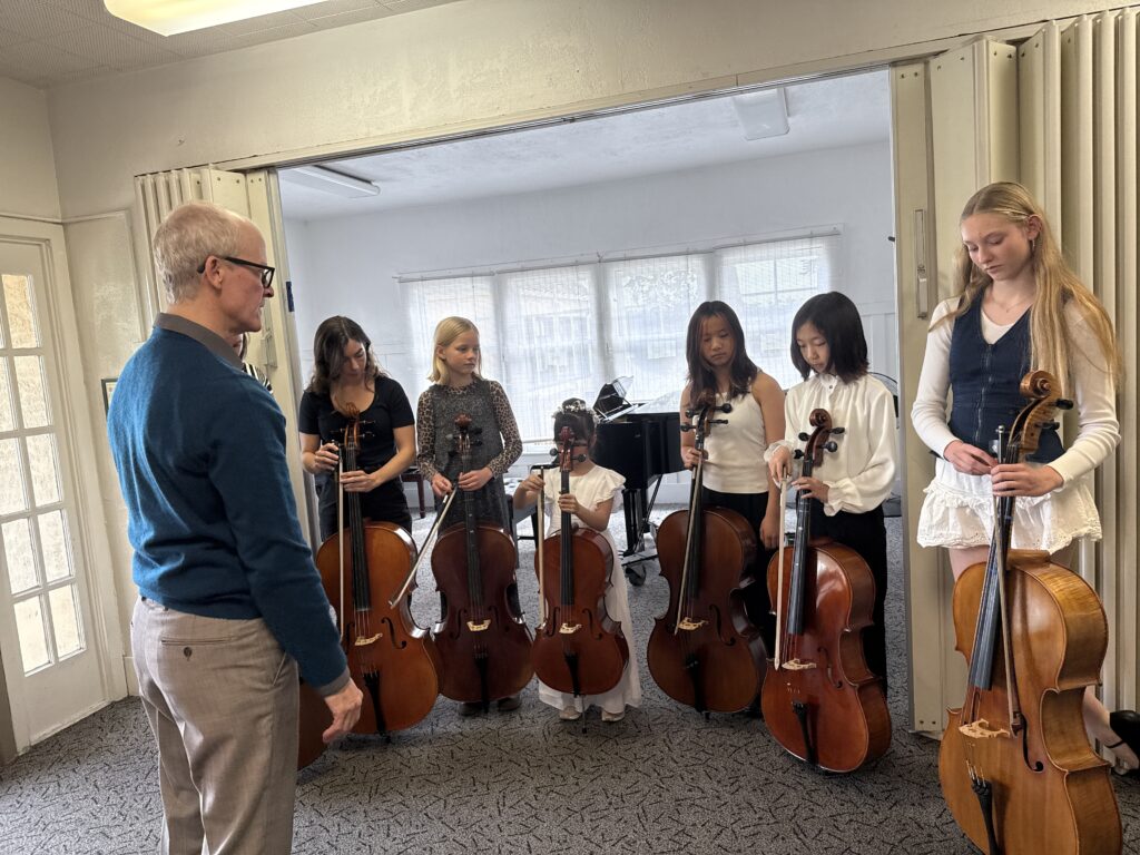 A music teacher guides a group of young girls holding cellos during a lesson in a bright, spacious room with a piano.