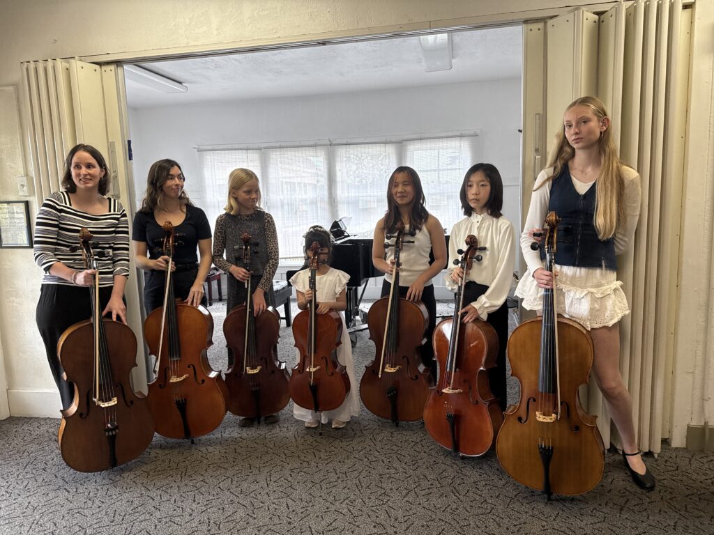 Seven young cellists, dressed in formal attire, stand together holding their cellos in a music studio, highlighting their dedication to performance.