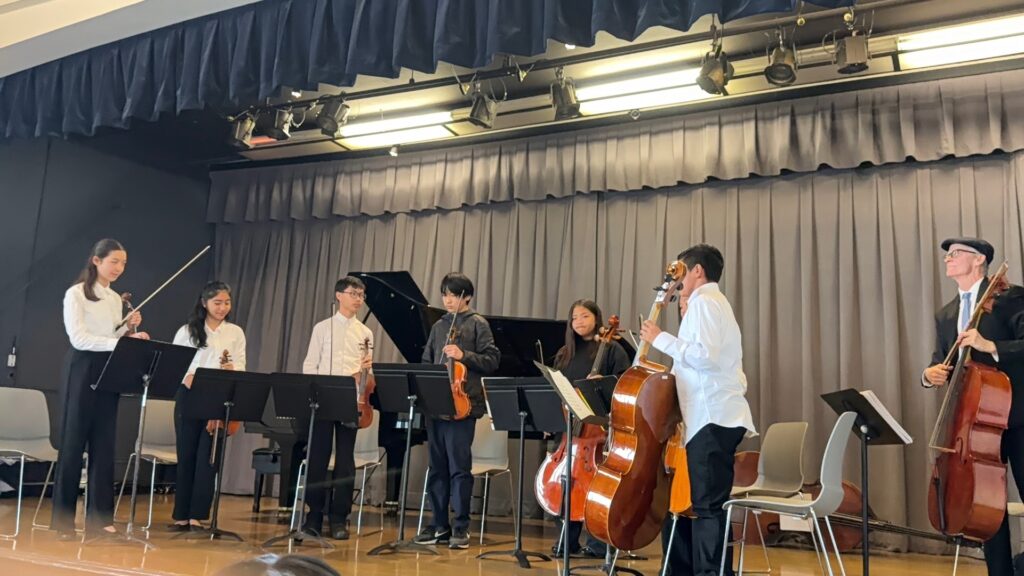 Seven musicians, including young students with violins and cellos and an instructor in a flat cap holding a cello, stand in front of music stands on a stage with a grand piano.